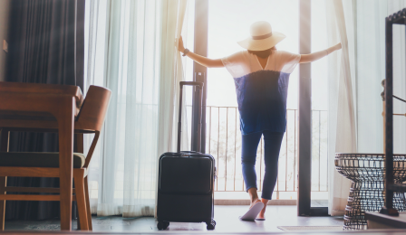 A woman with a sunhat on standing inside of a hotel room and looking out the window. She has a black suitcase next to her.