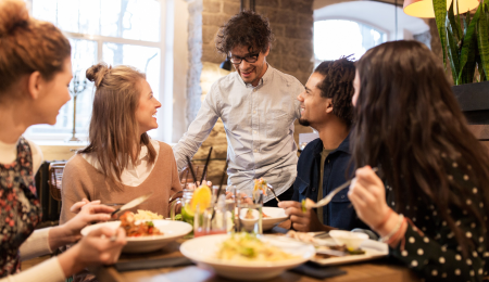 Four people smiling and eating food at a table in a restaurant, talking to a waiter.