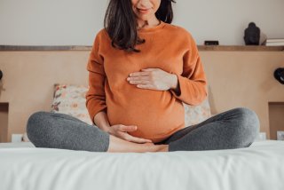 A pregnant person in sweatpants and a sweatshirt sits cross-legged on a bed.